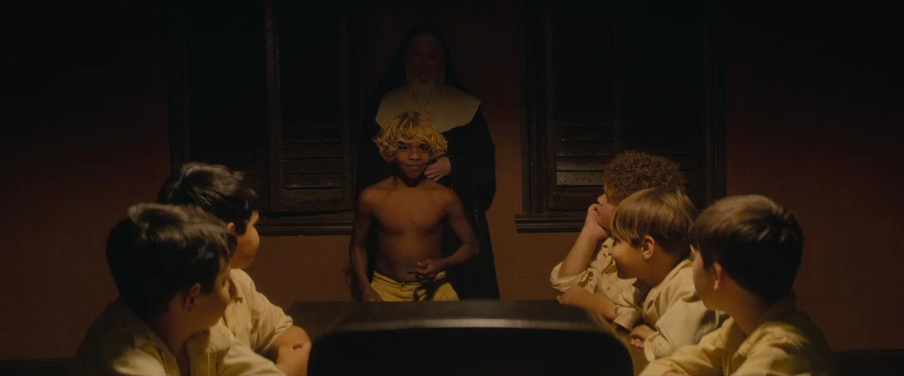 The New Boy (2023) movie still: medium — A dim room features a wooden table where several children in light shirts sit. A boy with blonde hair…