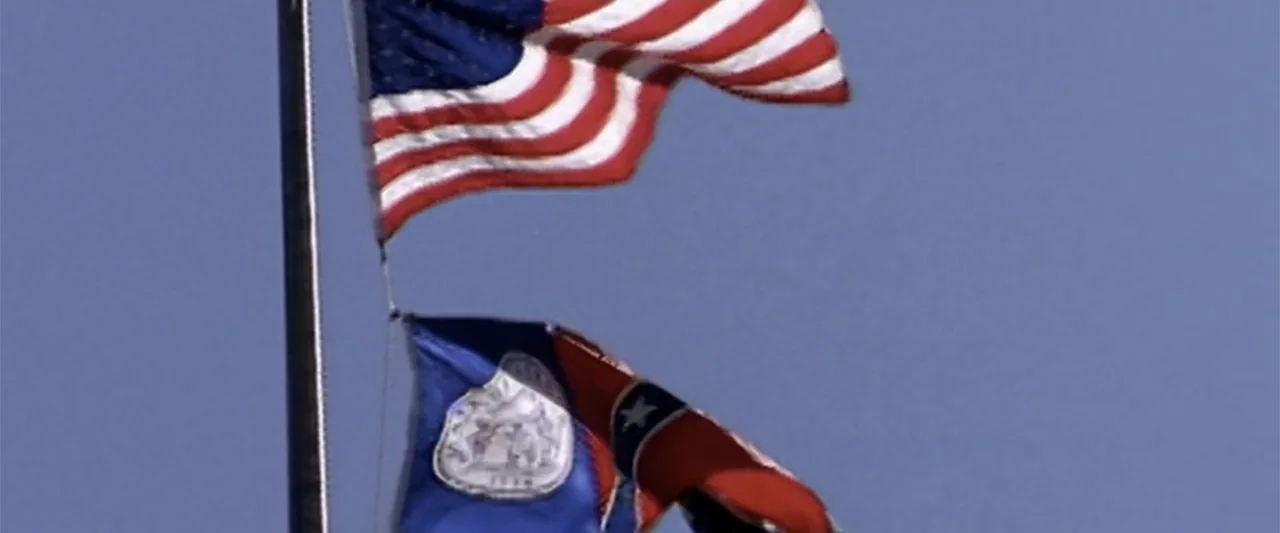 All In: The Fight for Democracy (2020) movie still: wide — Two flags fly from a metal pole against a solid blue sky background. The American flag is…