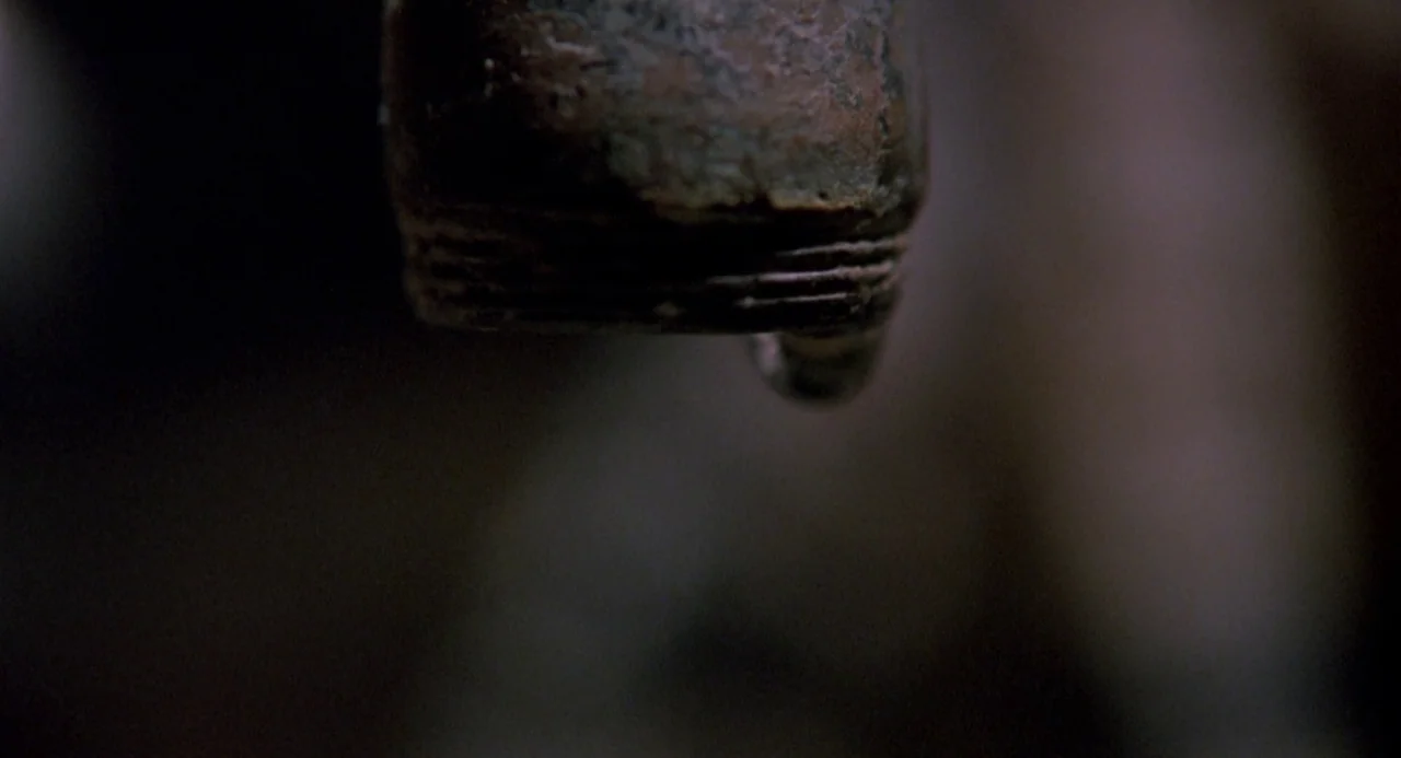 Boyz In The Hood (1991) movie still: closeup — A corroded metal pipe is centered in the dark, out of focus frame. A single clear water droplet hangs…