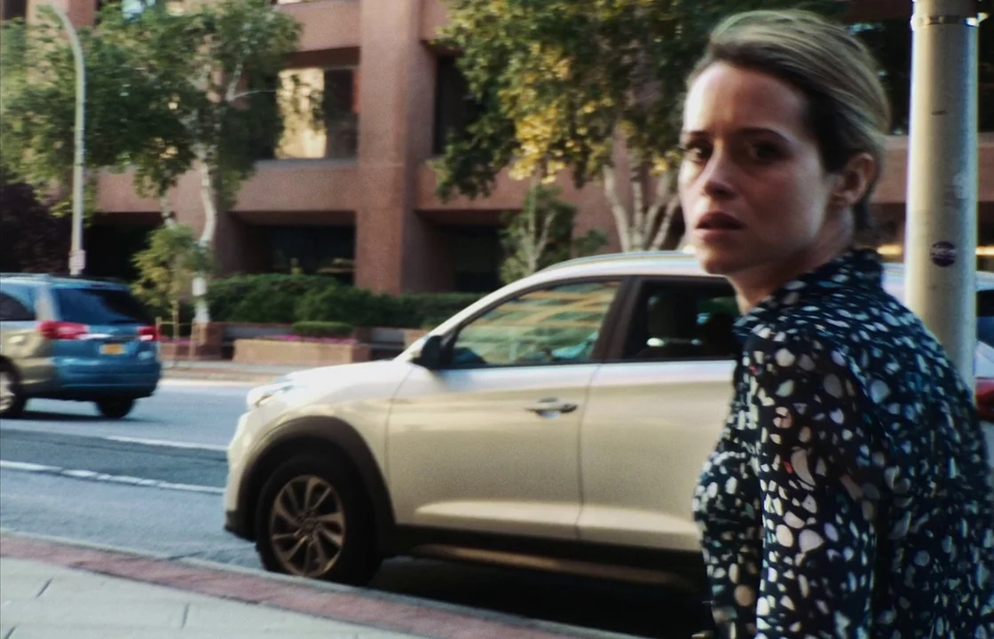 Unsane (2018) movie still: medium — A paved sidewalk runs alongside a busy multi-lane road lined with office buildings. A woman in a patterned…