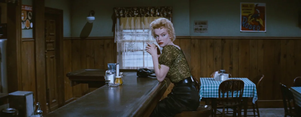 Bus Stop (1956) movie still: medium — A wood-paneled diner features a counter and tables covered in checkered tablecloths. A woman in a green lace…