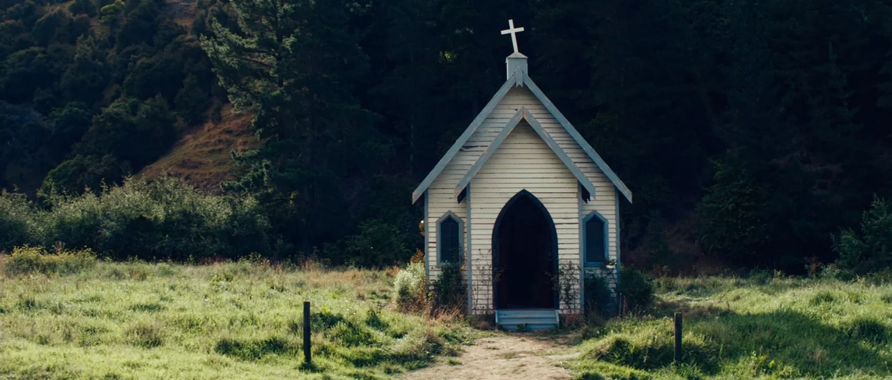 Z for Zachariah (2015) movie still: wide — A small weathered white wooden church sits in the center of a grassy field. Dense dark green trees and…