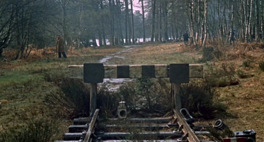 Fahrenheit 451 (1966) movie still: wide — A rural landscape features abandoned train tracks leading to a weathered timber stop barrier. Trees line…