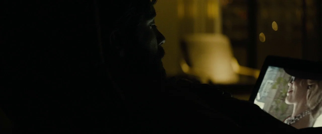 Enemy (2013) movie still: closeup — A man in a dark hoodie is seen in silhouette in a dim room. He watches a video playing on a small tablet screen…