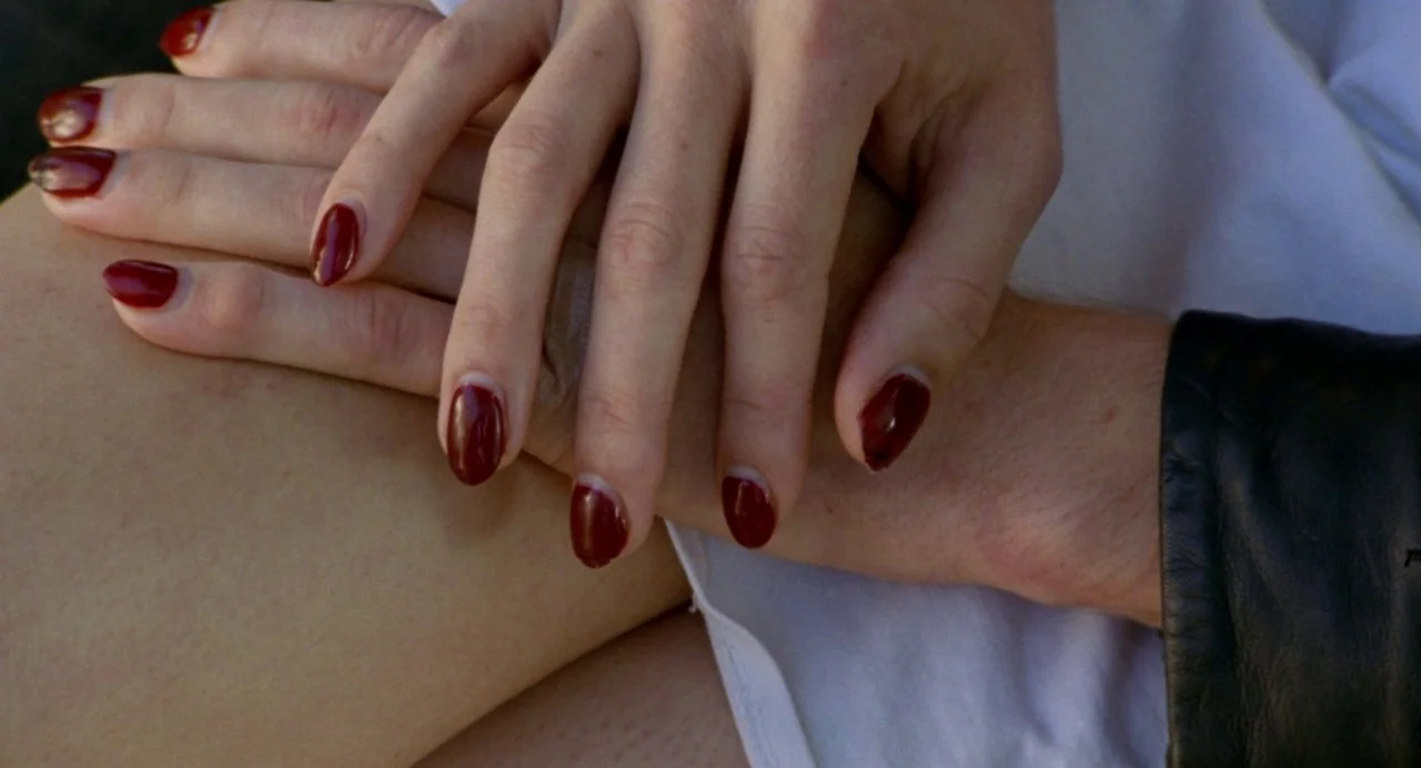 So Pretty (2019) movie still: closeup — Two hands with dark red manicured nails are clasped over a lap. A person wears white fabric and a black…