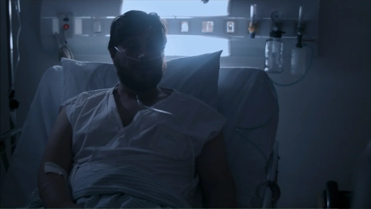 The Grotto (2022) movie still: medium — A hospital room at night is lit by dim, cool-toned ambient light. A bearded man lies in a bed wearing a…