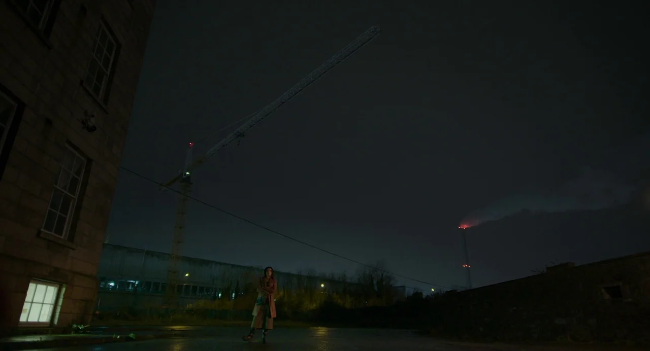 All You Need Is Death (2023) movie still: wide — A dark, paved industrial area under a night sky at the base of a crane. A solitary woman in a long…