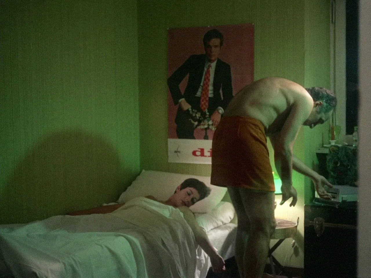 Dillinger is Dead (1969) movie still: medium — A small hotel room features lime green textured walls and a vintage travel poster. A shirtless man…