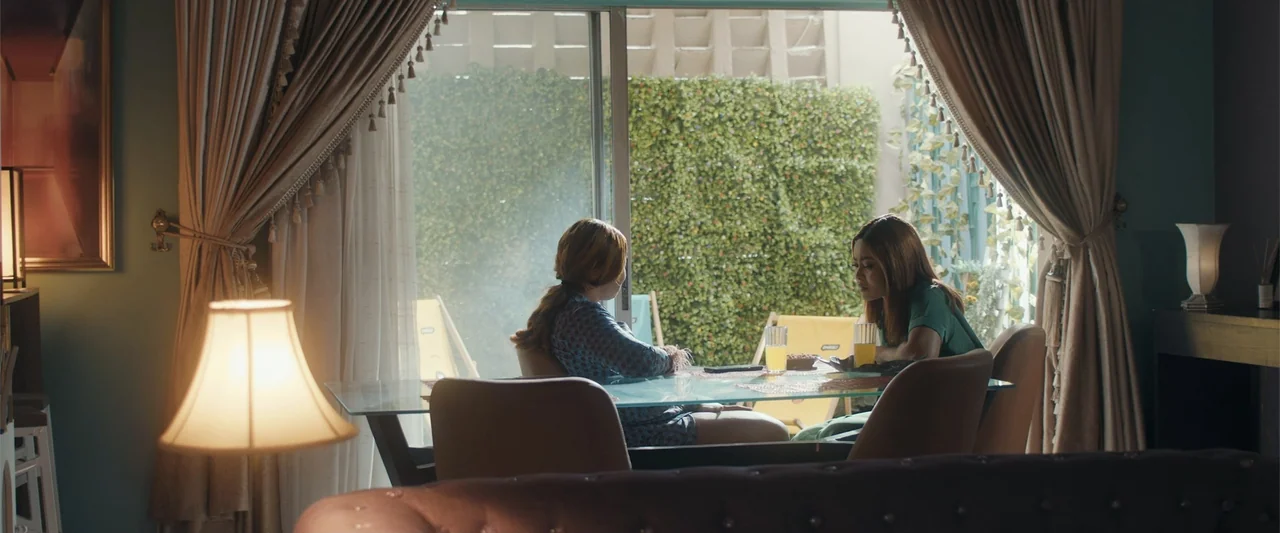 Big Love (2012) movie still: medium — A well-lit room features a glass table positioned in front of sliding glass doors. Two young women sit across…