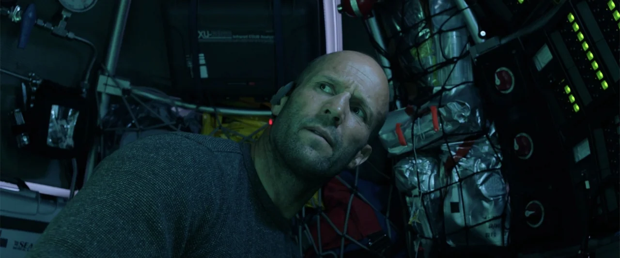 The Meg (2018) movie still: medium — A dark, cramped interior filled with complex technical gear and cargo netting. A man sits in a confined space…