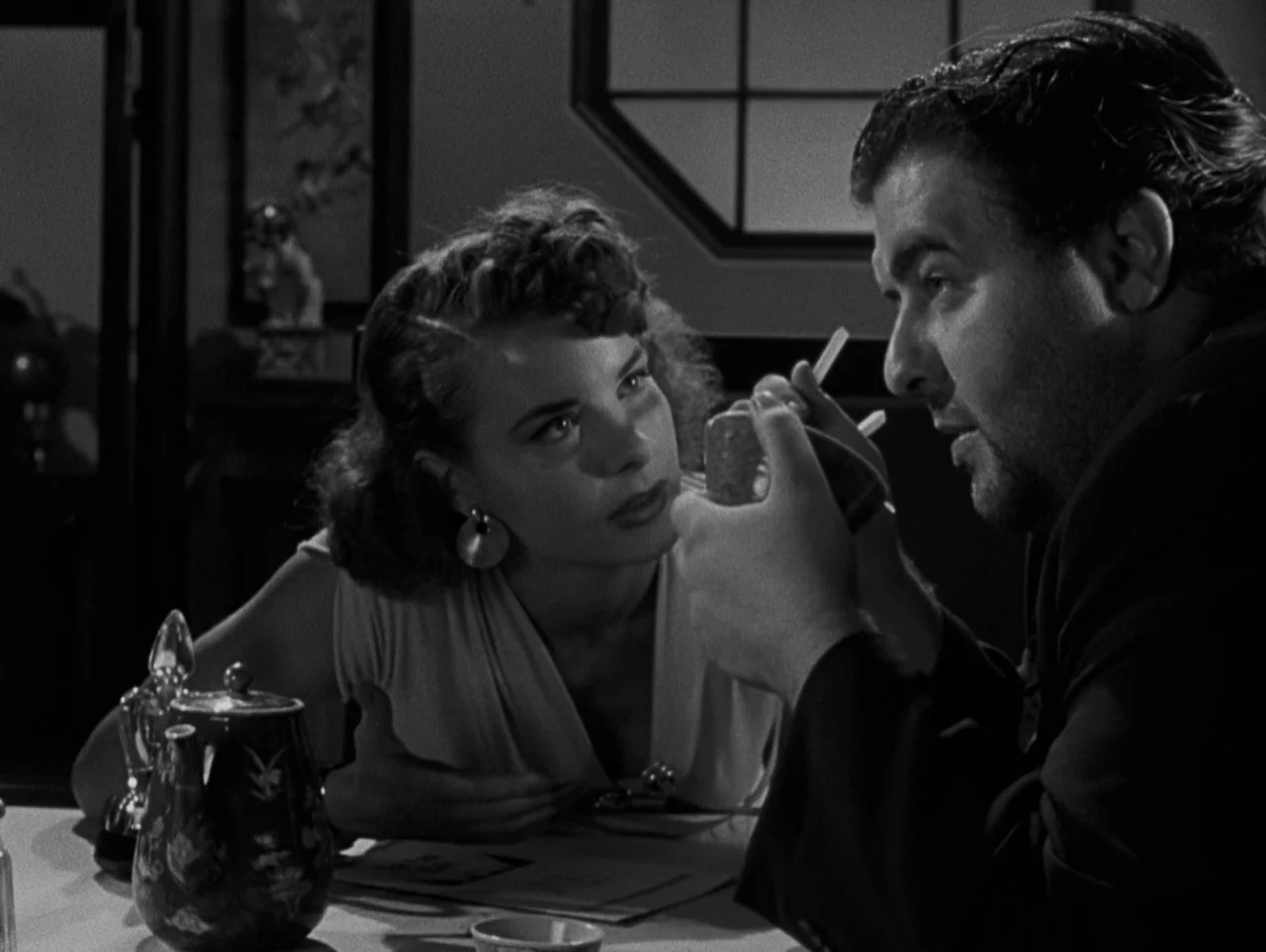 Pickup on South Street (1953) movie still: medium — A dark restaurant table features a teapot and a pair of white chopsticks. A woman watches a man…