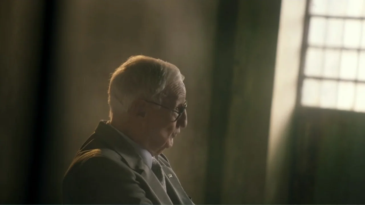 Transfers (2024) movie still: medium — An interior space with a tall, grid-patterned window casting light against a muted wall. An elderly man in a…