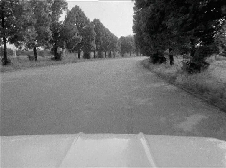 The American Soldier (1970) movie still: establishing — A rural paved road is lined with rows of mature trees on both sides. A vehicle drives along…