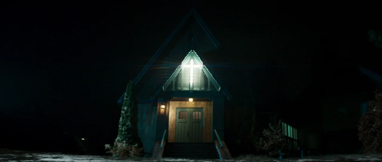 The Sacrifice Game (2023) movie still: establishing — A dark wooden church stands at night with a glowing cross above the door. Exterior steps lead…