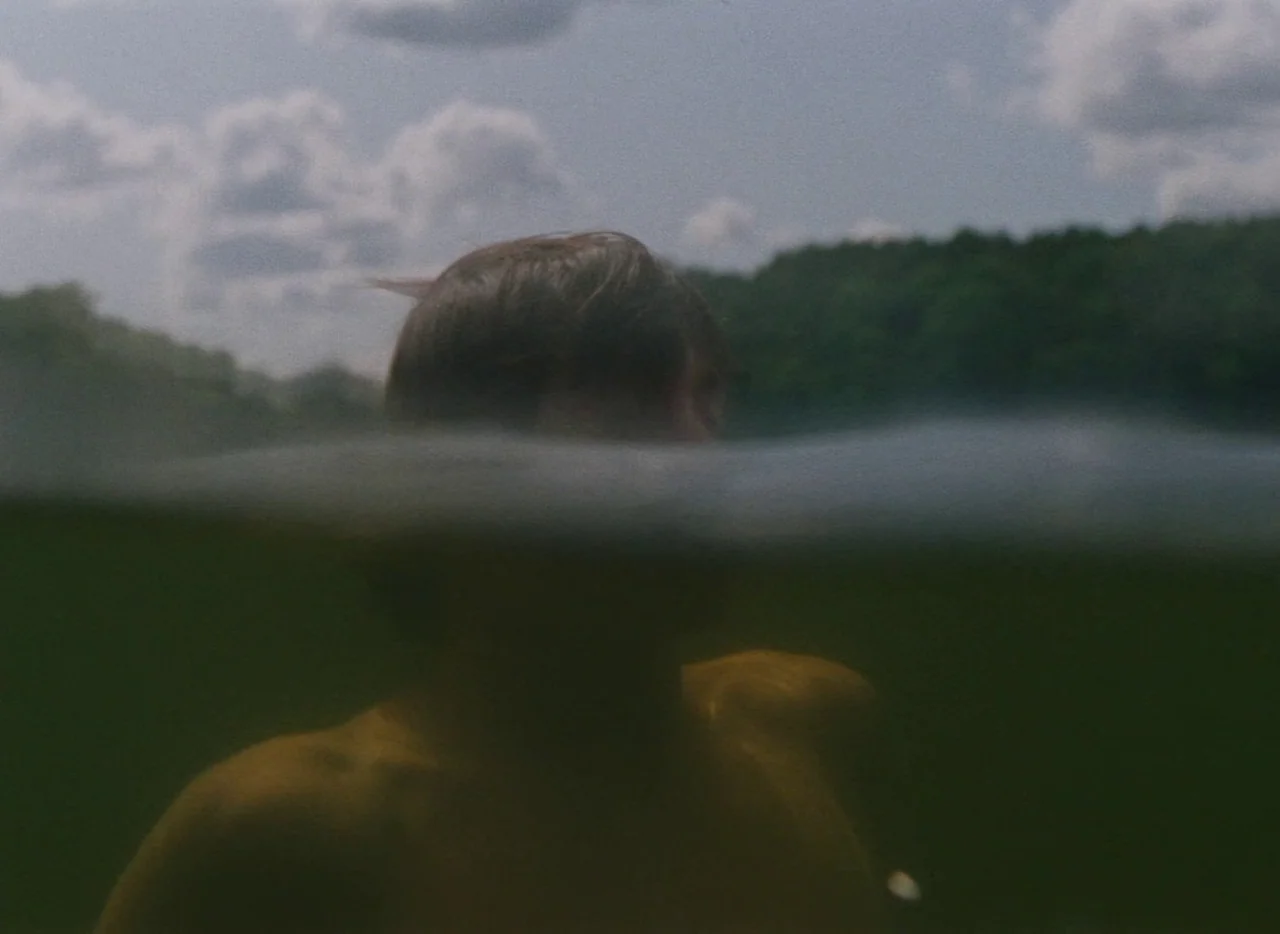 Falcon Lake (2022) movie still: medium — A split-level shot shows a man emerging from a murky, dark green lake. Clouds and a line of green trees…