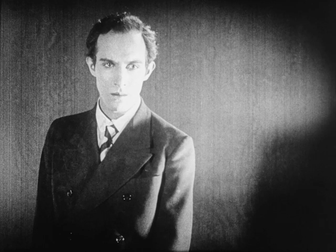 Un Chien Andalou (1929) movie still: medium — A man stands against a dark, textured wall in a minimalist space. He wears a formal suit and looks…
