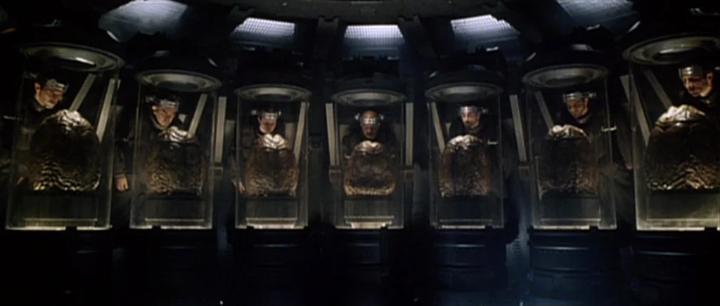 Alien: Resurrection (1997) movie still: wide — A dark, industrial room houses seven vertical glass pods arranged in a symmetrical row. Each pod…