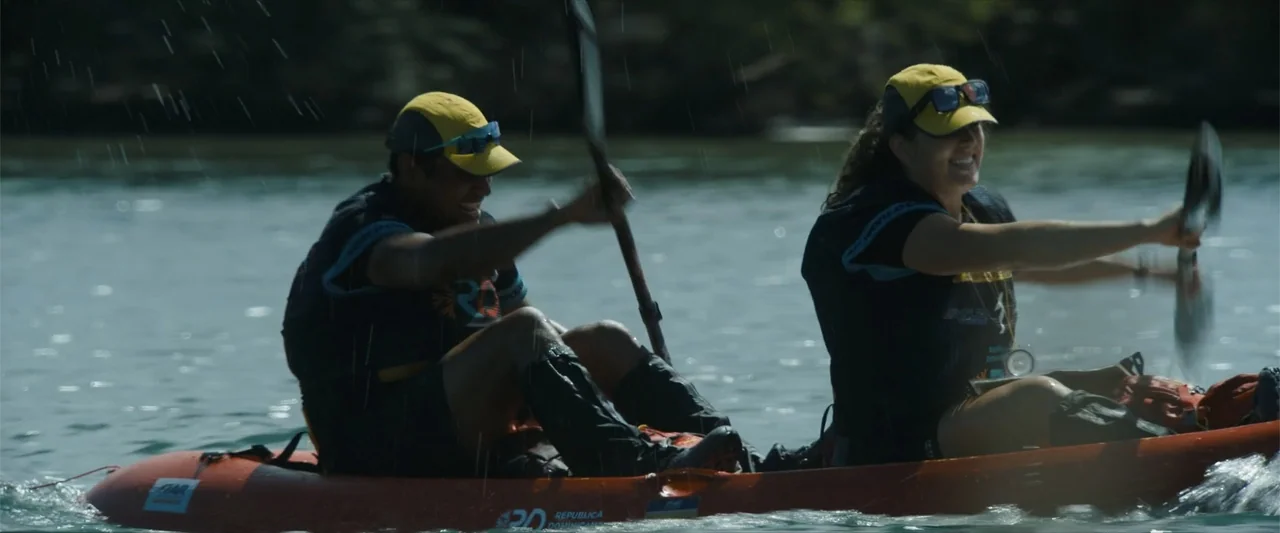 Arthur the King (2024) movie still: medium — A wide river scene featuring an orange kayak moving through open water. A man and woman in athletic…