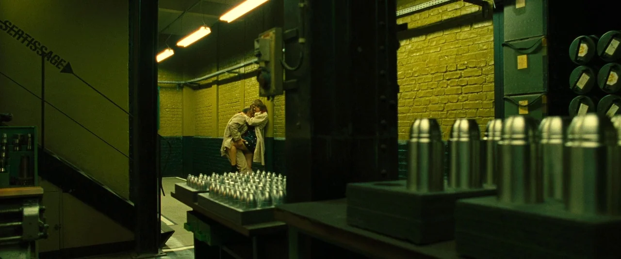 Micmacs (2009) movie still: wide — A dimly lit industrial room features brick walls and rows of machined metal components. A man and woman are…