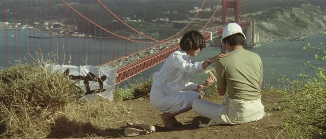 Take Me Away! (1978) movie still: wide — A grassy hillside overlook features the Golden Gate Bridge in the background. Two individuals crouch on the…