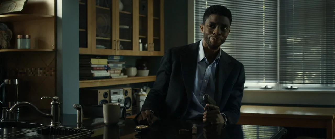 21 Bridges (2019) movie still: medium — A modern residential space with dark countertops and wooden cabinetry visible in the background. A man holds…