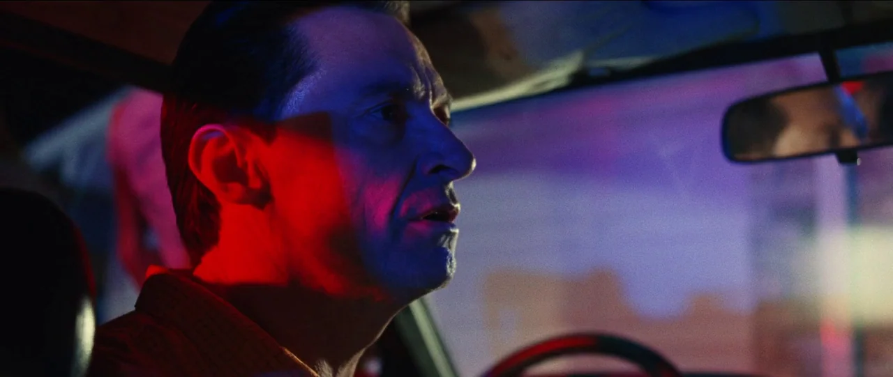 Bad Education (2019) movie still: closeup — A man drives a car at night illuminated by harsh colored exterior neon lights. The interior of the…