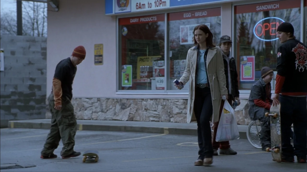 Final Destination 2 (2003) movie still: fullbody — A convenience store exterior with glass windows and an open sign in late afternoon. A young woman…