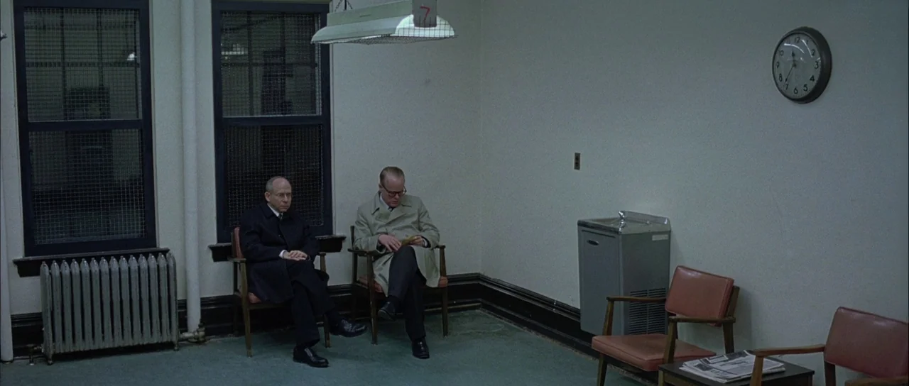 Capote (2005) movie still: wide — A sparse, institutional room features two men sitting on chairs near a radiator. The setting includes two barred…
