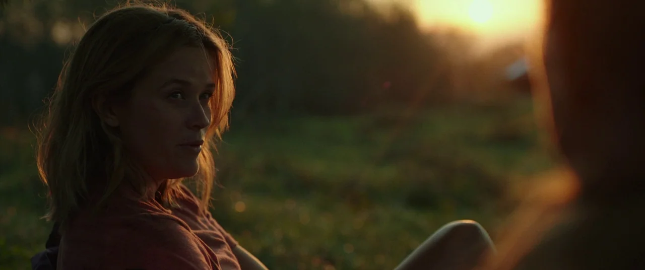 Wild (2014) movie still: medium — A grassy field is illuminated by the low angle light of the sunset. A woman sits in the foreground looking toward…