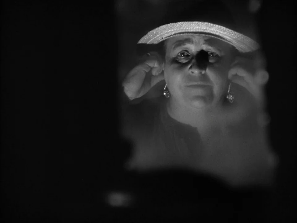 The Grapes of Wrath (1940) movie still: closeup — The frame depicts a woman reflected in a mirror inside a dimly lit space. She is adjusting her…