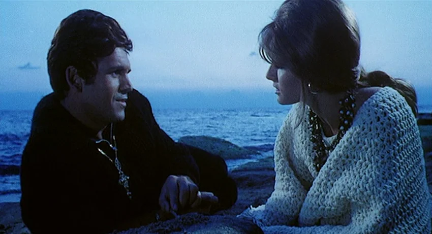 5 Dolls for an August Moon (1970) movie still: medium — A man and woman kneel on a rocky beach at twilight near the water. They face each other as…