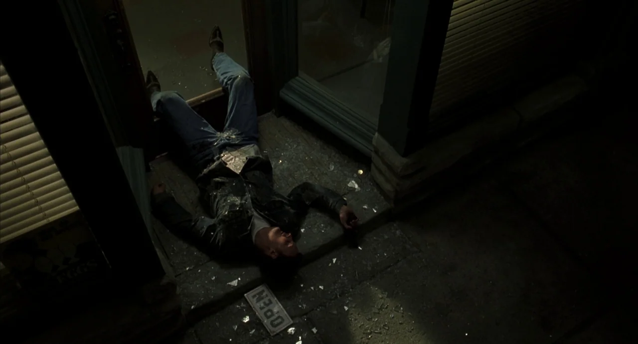 A History of Violence (2005) movie still: wide — A man lies on the concrete threshold in front of a shattered glass door. Broken glass fragments are…
