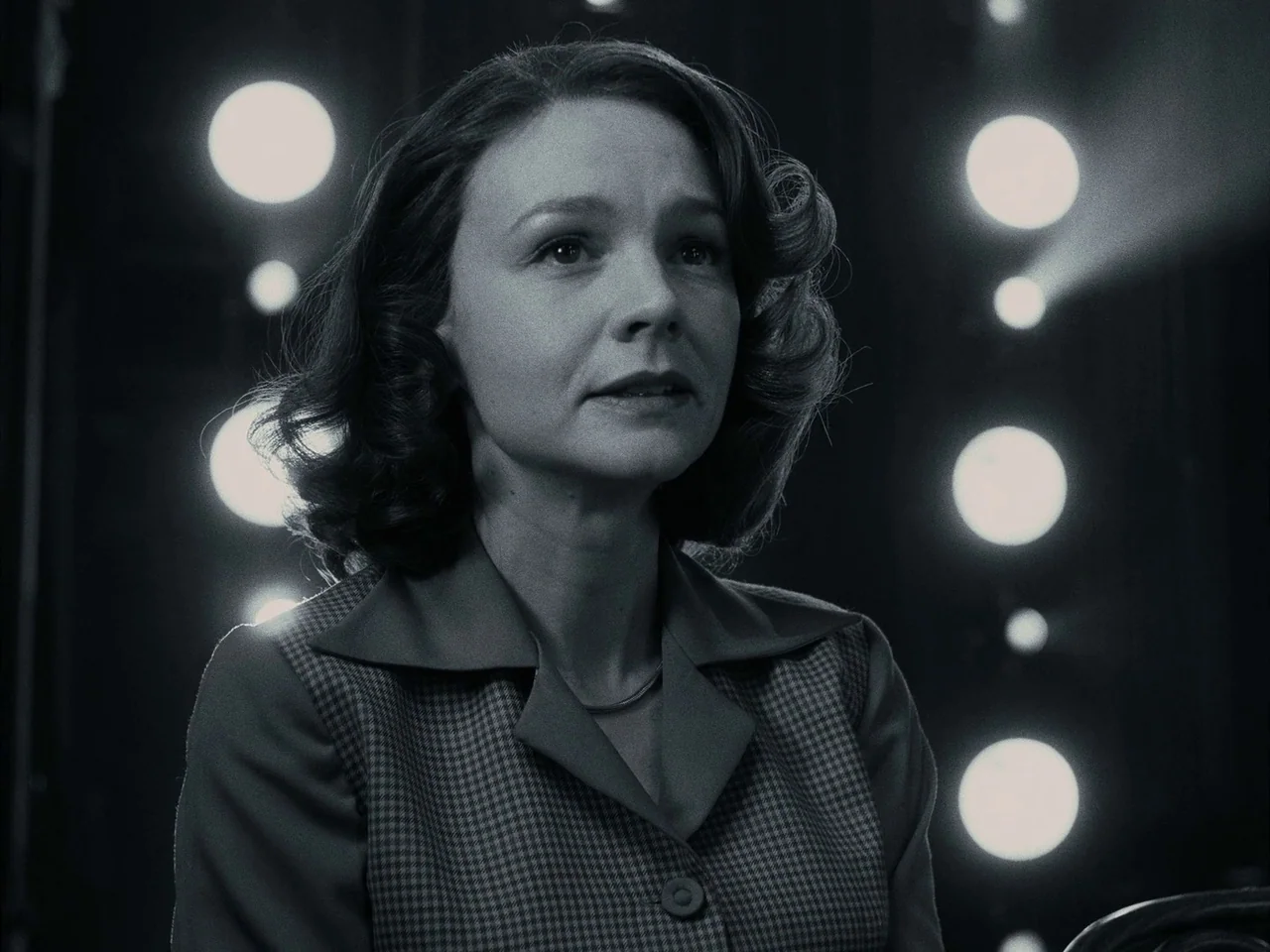 Maestro (2023) movie still: medium — A woman stands in front of a dark backdrop featuring arranged circular stage light fixtures. She looks towards…