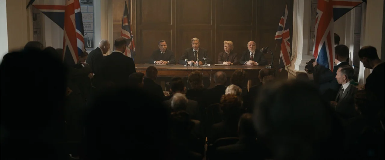 Sardar Udham (2021) movie still: wide — A formal wooden room featuring four officials seated at a long table with microphones. Several British flags…