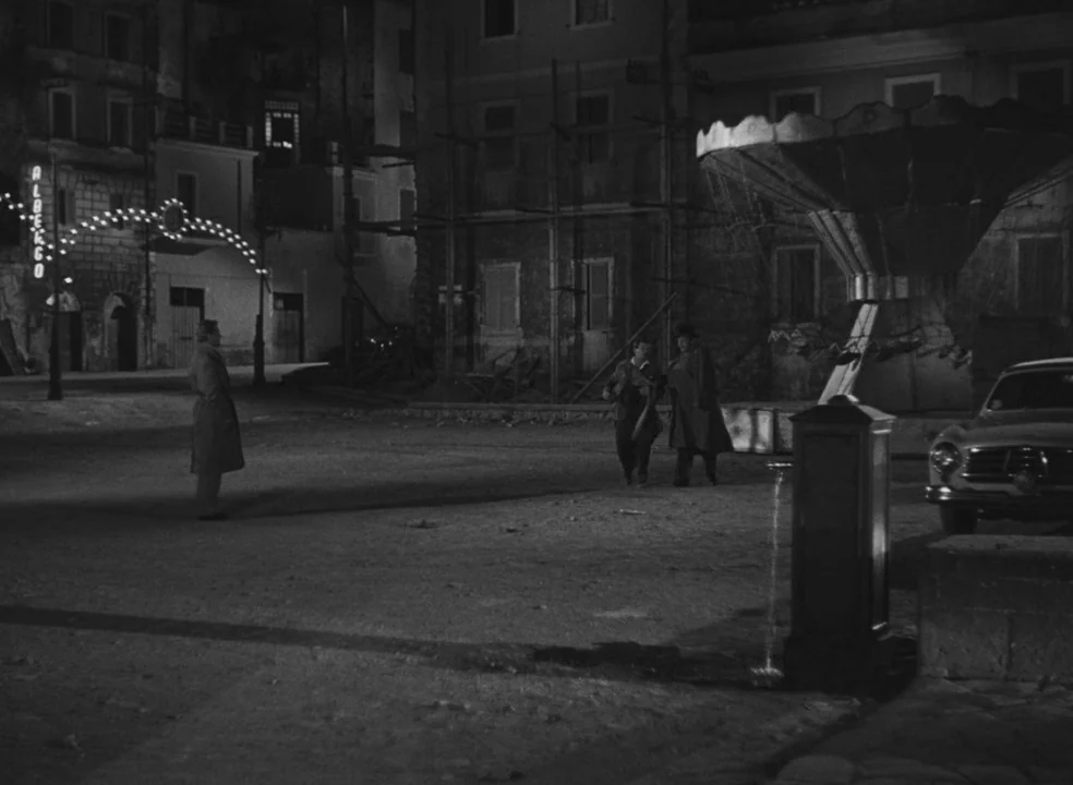 Il Bidone (1955) movie still: wide — An empty cobblestone square at night with scaffolding on buildings and a water fountain. Three men walk across…