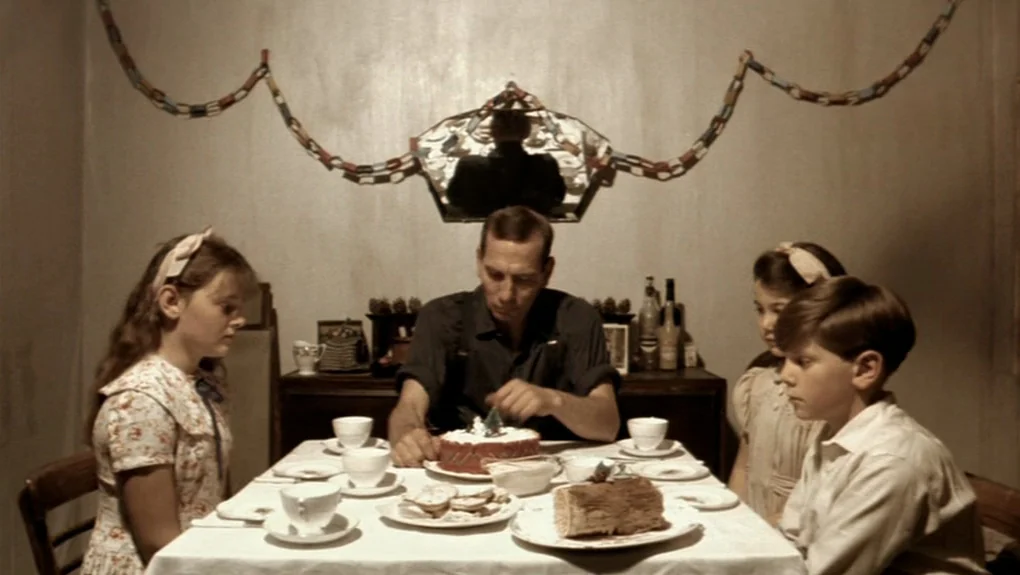Distant Voices, Still Lives (1988) movie still: medium — A 1940s-style room features paper chains hanging against a plain wall above a buffet. A man…