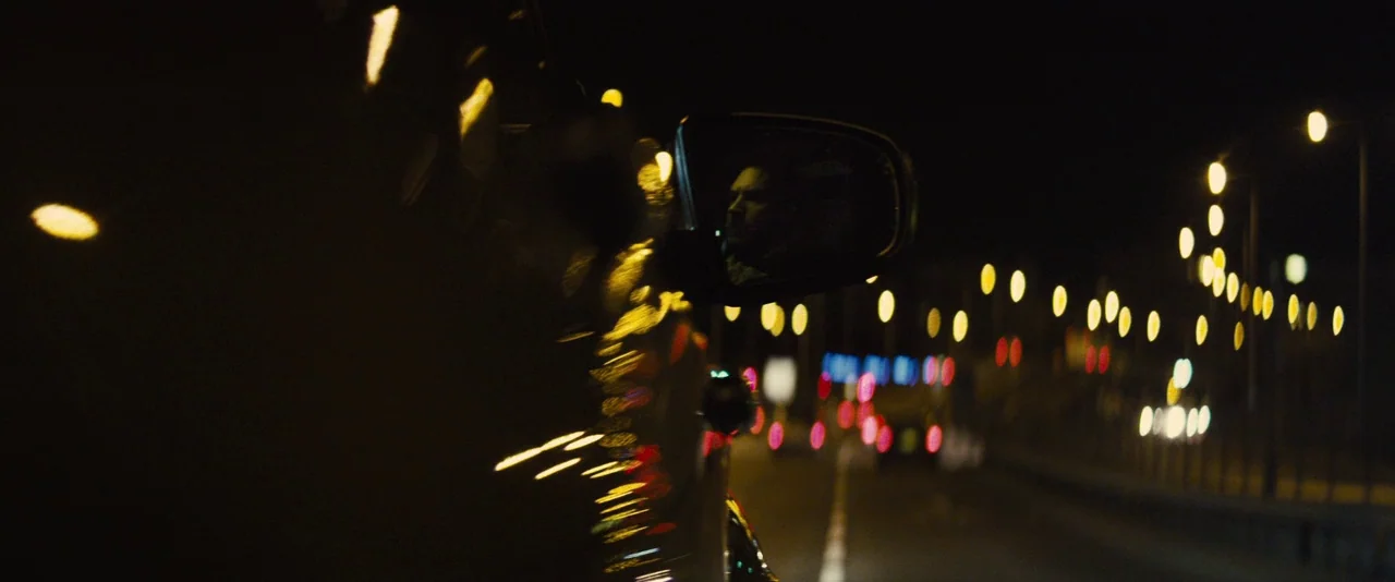 Locke (2013) movie still: closeup — A night view of a highway reflected in a moving vehicle's side-view mirror. The driver is visible within the…