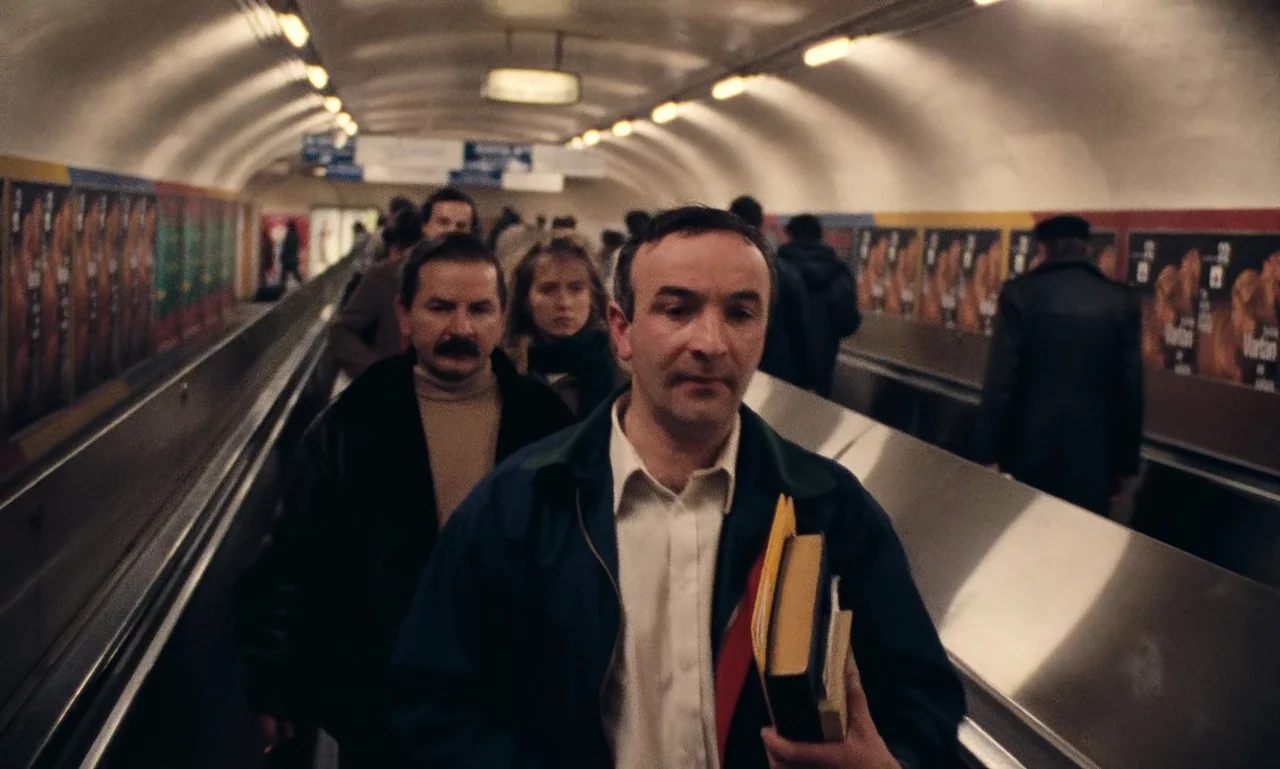 A Tale of Winter (1992) movie still: medium — A crowded subway terminal with a long escalator tunnel lined with advertisements. A middle-aged man…