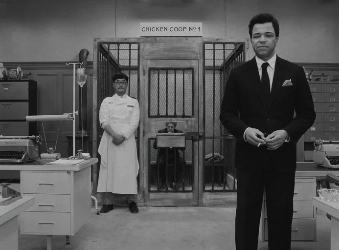 The French Dispatch (2021) movie still: fullbody — An interior space with a sign reading 'CHICKEN COOP NO 1' above a wire cage. Three men in…