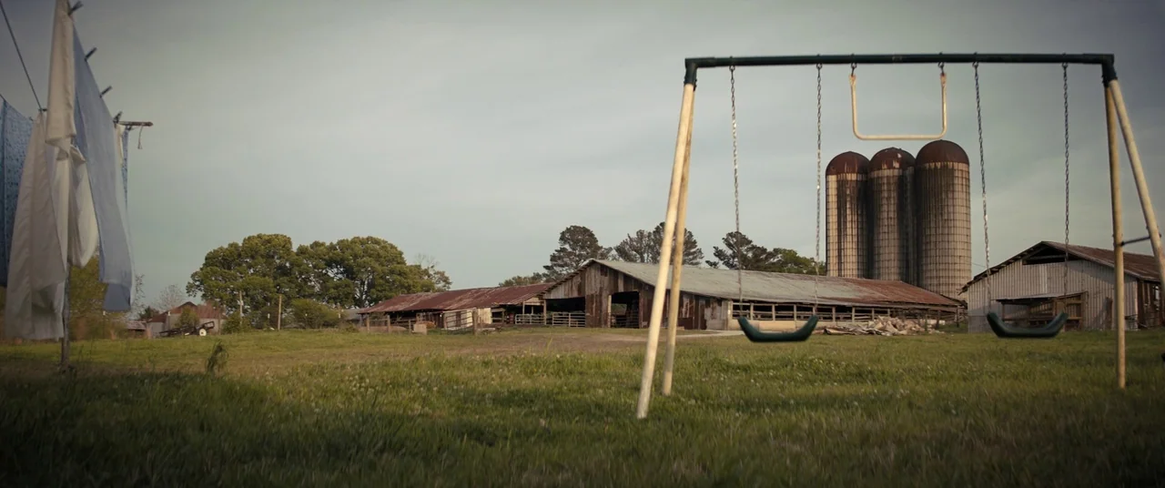 Brightburn (2019) movie still: wide — An open grassy farm field with a metal swing set and hanging laundry clotheslines. Three tall concrete silos…