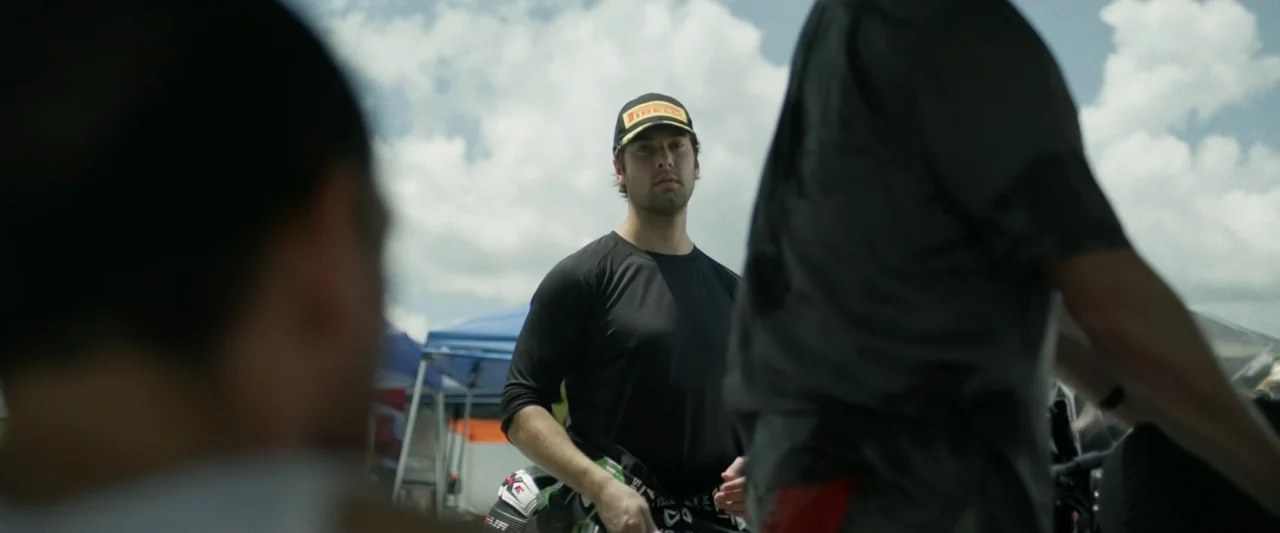 One Fast Move (2024) movie still: medium — A sunny outdoor paddock area features blurred tents and white clouds in the background. A young man in a…