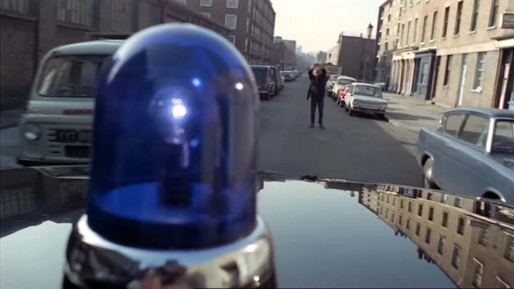 The Sorcerers (1967) movie still: wide — A residential street in London is lined with parked 1960s-era automobiles and tall brick buildings. A man…