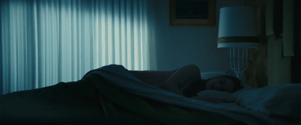 I'm Your Woman (2020) movie still: medium — A dimly lit bedroom features a bed positioned against a wall with sheer curtains. A woman sleeps under…