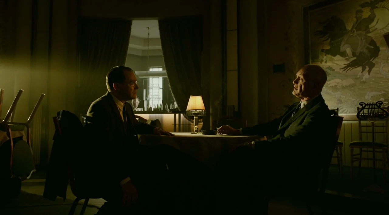 The Shape of Water (2017) movie still: medium — A dimly lit room features a small table with a lamp and two men seated. A large framed illustration…