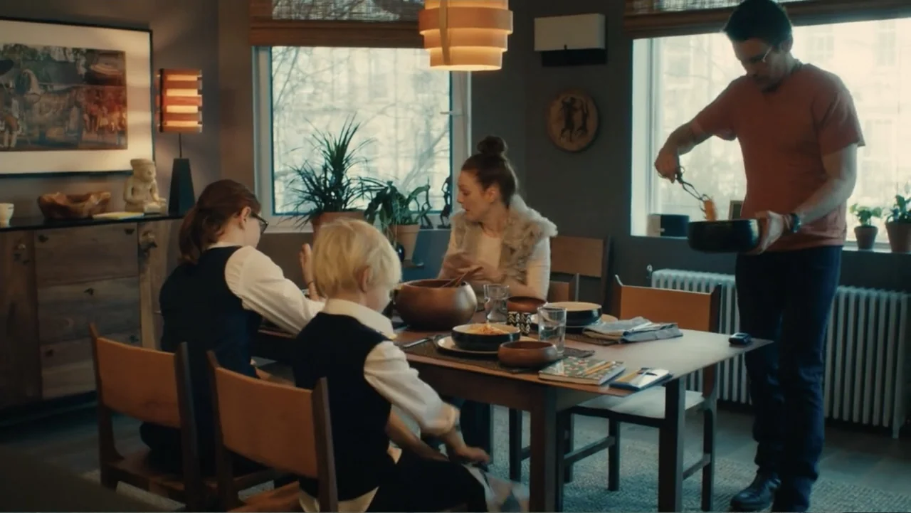 Maggie's Plan (2016) movie still: medium — A sunlit dining room features dark walls, wooden furniture, and various indoor house plants. A family is…