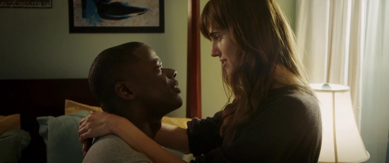 Get Out (2017) movie still: medium — A residential bedroom features a bed with blue and yellow pillows against teal walls. A man and woman sit on…