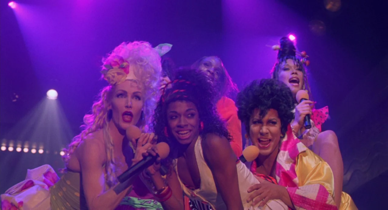 The Birdcage (1996) movie still: medium — A group of performers on a dimly lit stage during a musical performance. They are singing into handheld…