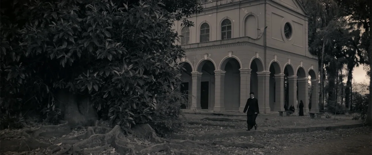I Trust in You (2016) movie still: wide — A historic building with an arched portico sits among mature trees and leaf-covered ground. A person…