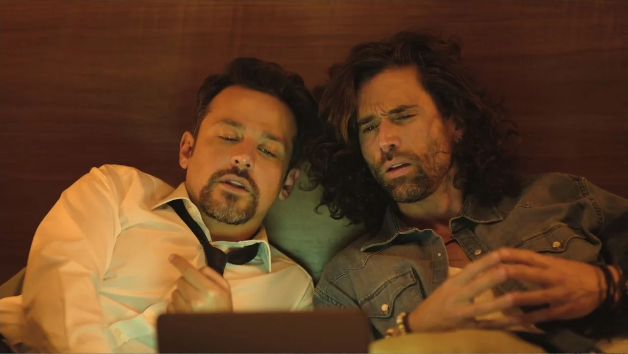 Double Pregnant 2 (2022) movie still: medium — Two men lie on a bed in a darkened room with wood-paneled walls. They watch the screen of a laptop…