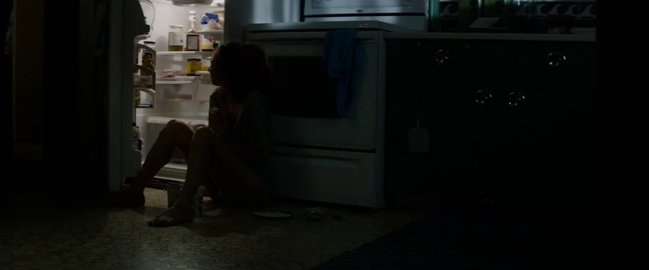 The Monster (2016) movie still: medium — A dark, residential space with a white oven next to an open refrigerator. A woman sits on the patterned…